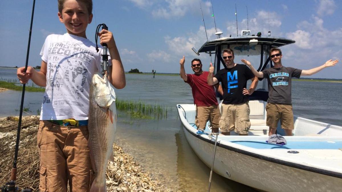 Boy holding up fish on beach with people in boat in the background