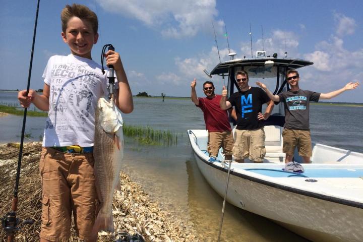 Boy holding up fish on beach with people in boat in the background