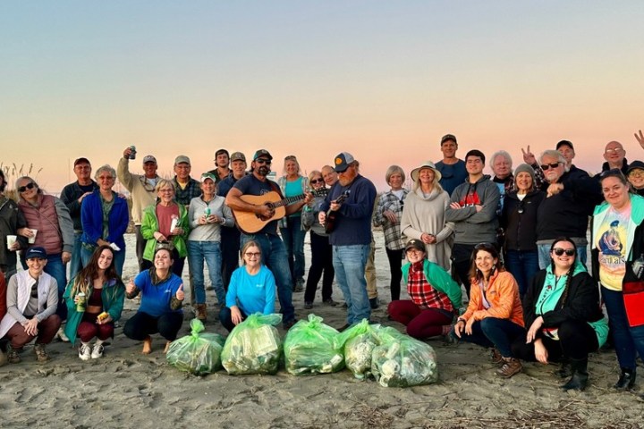 Group of people posing on a beach with trash bags and a guitar at sunset.