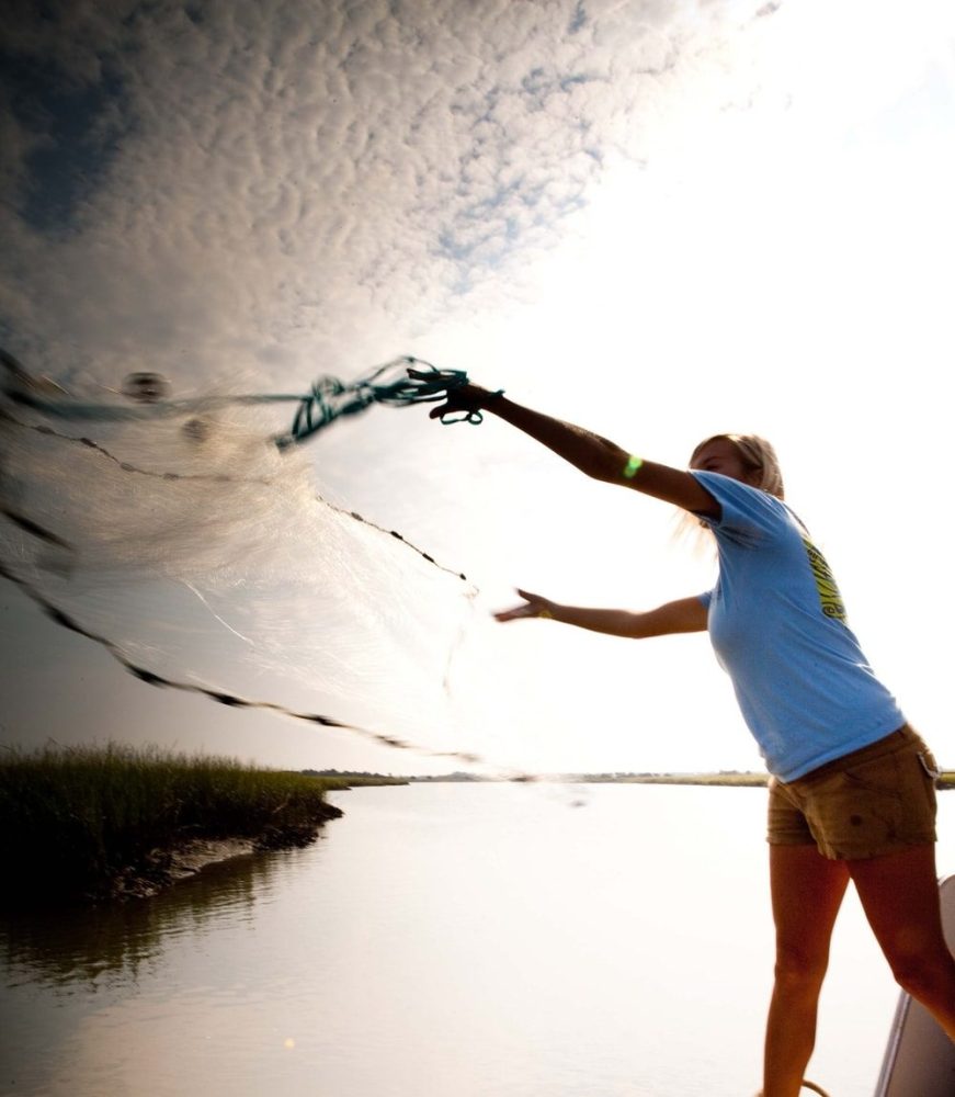 Person on dock casting a net into a river under a cloudy sky.