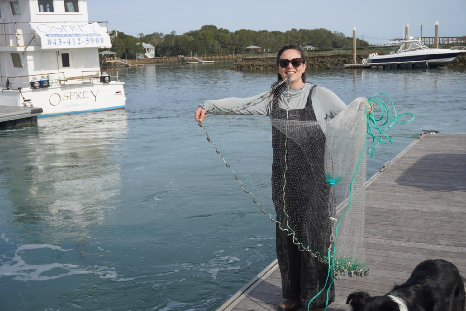 Person on dock holding a fishing net, with a boat named Osprey in the water and a dog nearby.