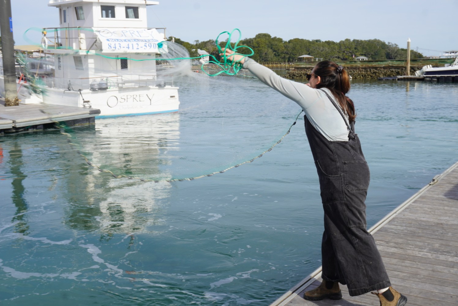 Person casting a fishing net into a harbor near a boat and dock.