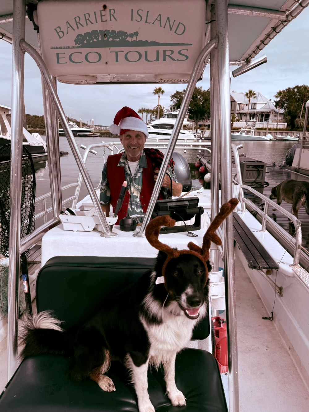 Dog with reindeer antlers on boat with man in Santa hat, 'Barrier Island Eco Tours' sign above.