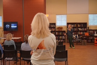 Audience member watches a speaker in a library setting with bookshelves and a presentation screen.